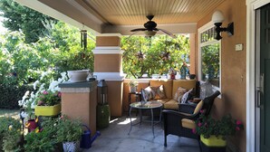Un patio al aire libre con un ventilador de techo, una zona de estar con un sofá y sillones, y diversas plantas en macetas.
