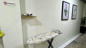 Un corridoio con un cartello di divieto di fumo, uno scaffale con tazze, un asse da stiro con motivi floreali e due stampe botaniche incorniciate al muro.