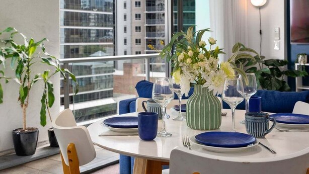 A dining area with a round table, chairs, a vase of flowers, and a view of buildings through large windows.
