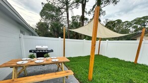 A wooden picnic table with plates and a grill in a backyard with a white fence and grass. - NEW Gorgeous Sunshine-Up 6 Guests-Private Backyard (Port Orange)