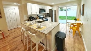A compact kitchen with white cabinets, a microwave, and a refrigerator. There is a dining area with a table and chairs, and a view of a backyard through a sliding glass door. - NEW Gorgeous Sunshine-Up 6 Guests-Private Backyard (Port Orange)