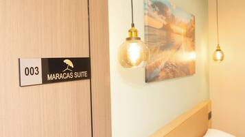 Habitación de hotel con puerta de madera, una señal que indica el número de habitación 003 y la etiqueta Maracas Suite. Hay dos lámparas colgantes y un cuadro en la pared.