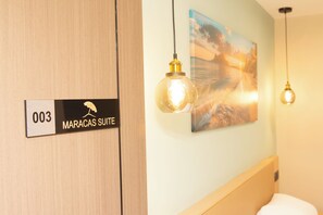 A hotel room with a wooden door, a sign indicating room number 003, and a Maracas Suite label. There are two hanging light fixtures and a painting on the wall. - Tstays Smart Hotel (Port of Spain)