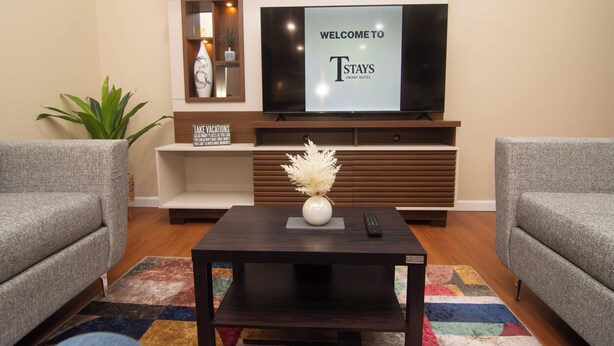 A modern living room with a flat-screen TV, two grey sofas, a dark coffee table with a vase, and a colorful rug. - Tstays Smart Hotel (Port of Spain)