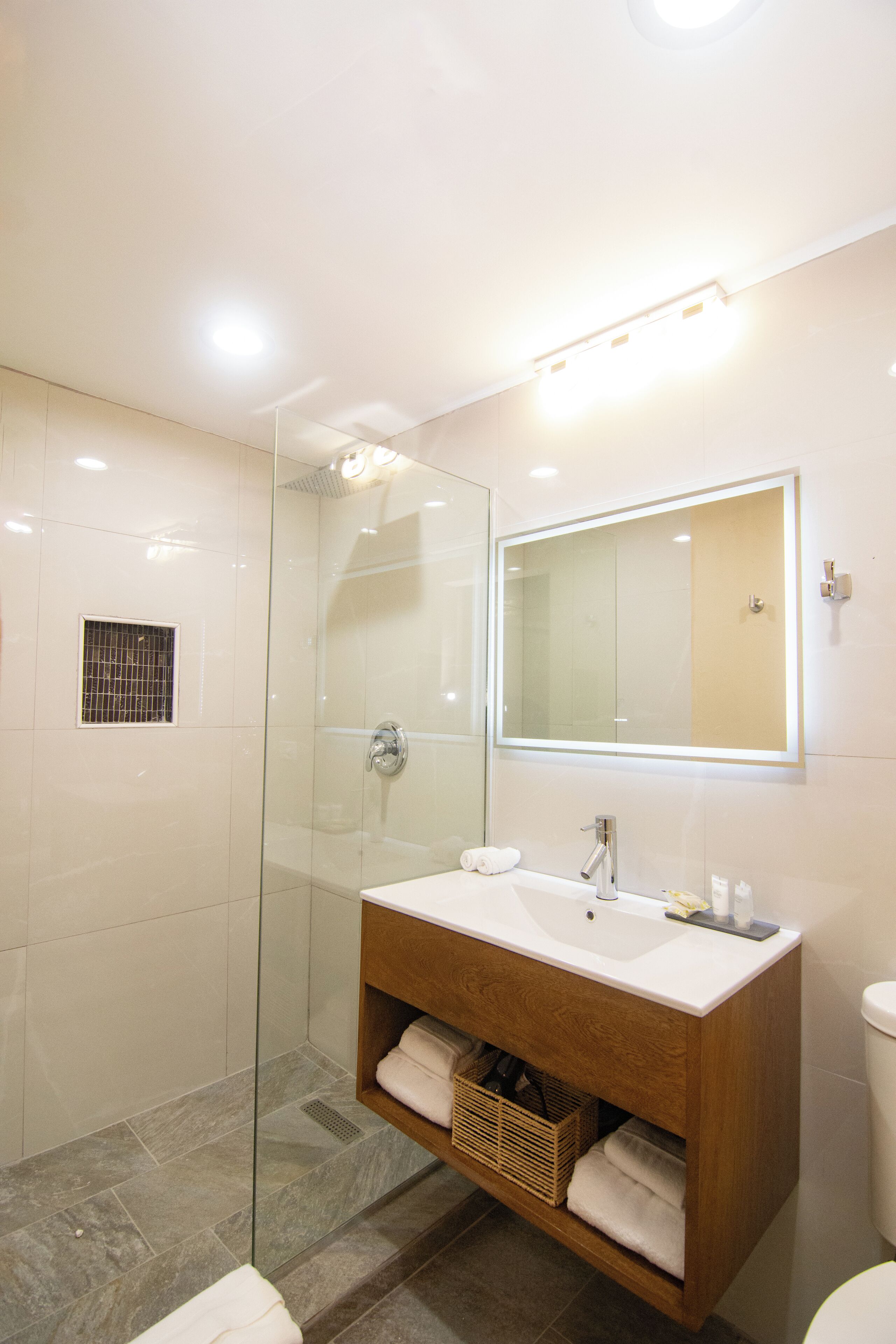 Un baño moderno con ducha envuelta en vidrio, tocador de madera con lavamanos y un espejo grande.