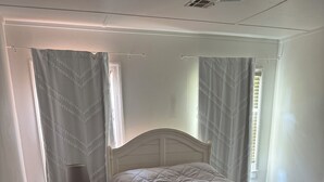 Ein Schlafzimmer mit Holzboden, einem weißen Bett mit Kopfteil, einem Nachttisch mit Lampe und einem Fenster mit Vorhängen.