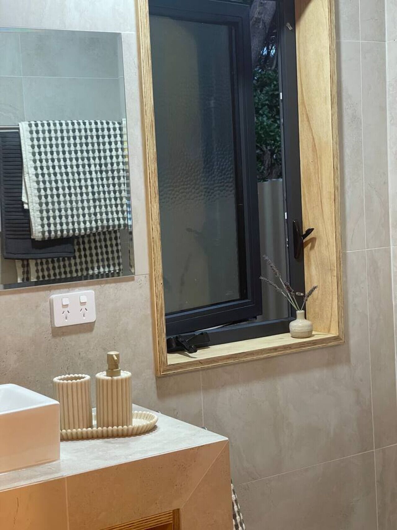 A bathroom with a mirror, a window with a wooden frame, a sink with a vase of dried plants, and a towel rack with a checkered towel.