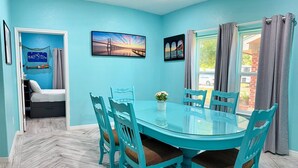 A dining area with a glass table and turquoise chairs, a painting on the wall, and a view through the window. - Coastal Comfort Retreat | Near Beach | Sleeps 10! (Rockport)