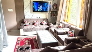 A modern living room with a sectional sofa, a coffee table, and a TV mounted on a marble wall. - Tranquil trails Naddi Mountain view (Dharamshala)