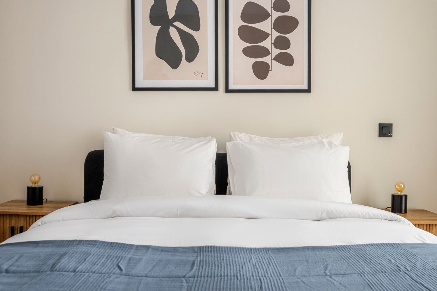 A neatly made double bed with white linens, two bedside tables with lamps, and two framed abstract art pieces on the wall.