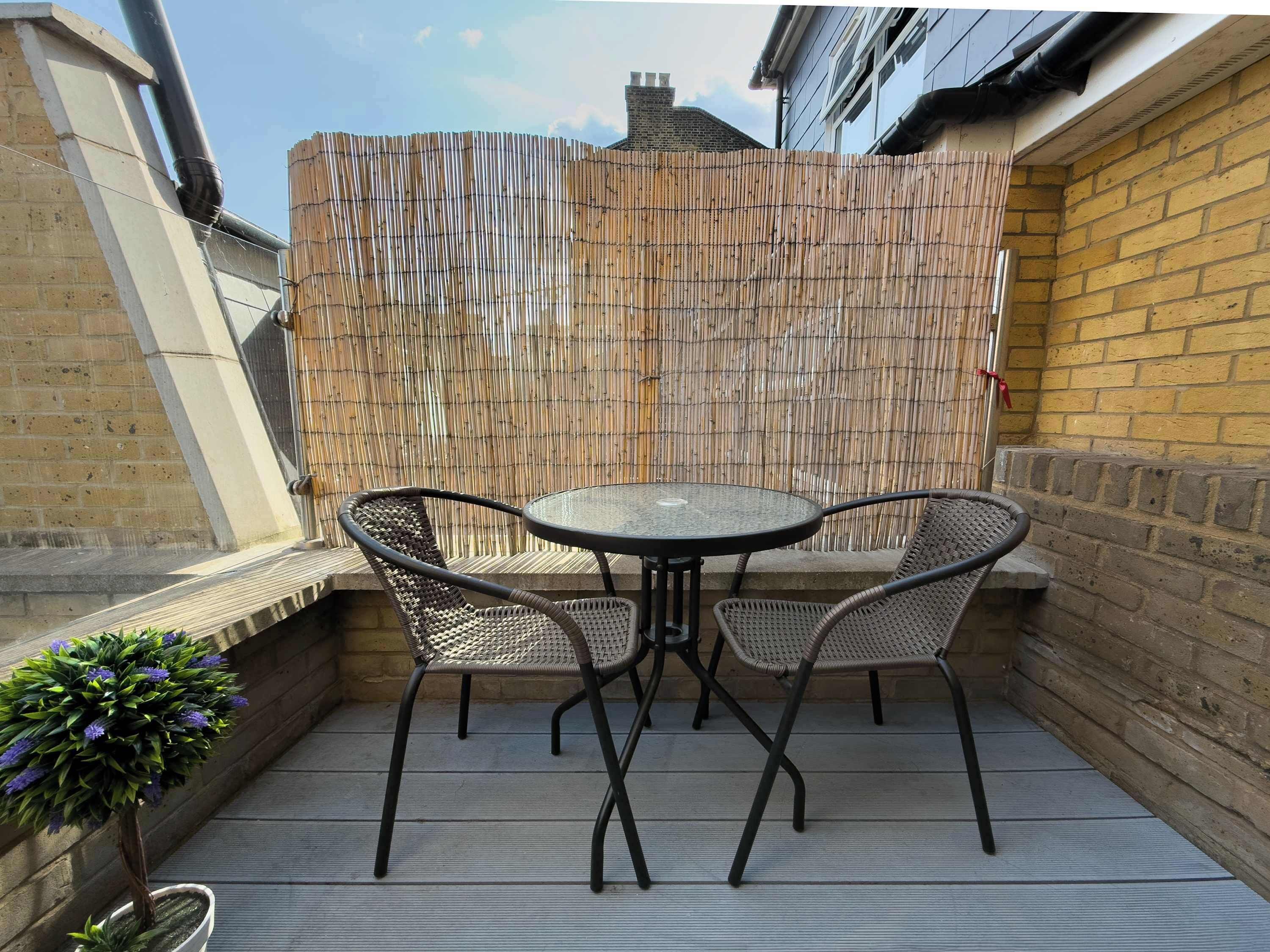 An outdoor patio with a round table and two chairs, a bamboo fence, and a potted plant.