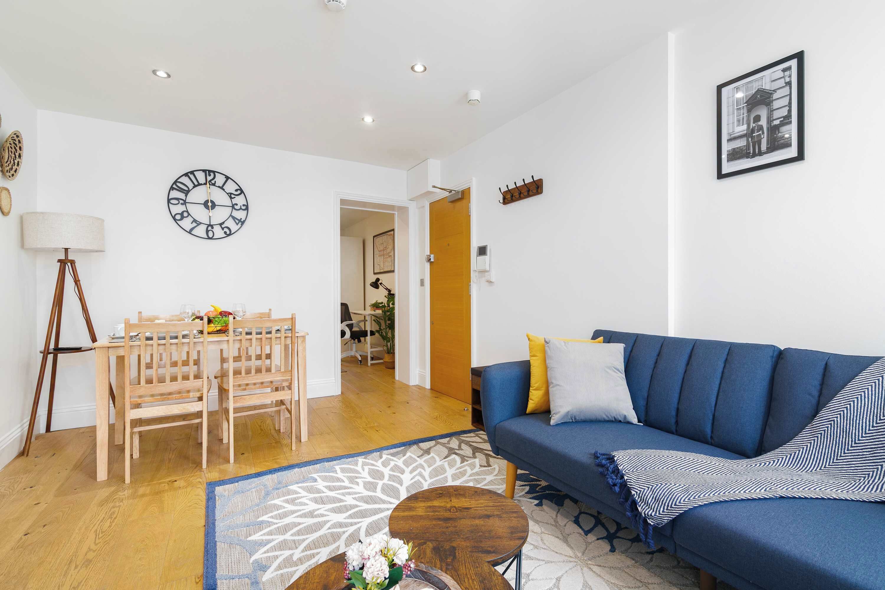 A living room with a blue sofa, a wooden dining table with chairs, a round coffee table, and a wall clock.