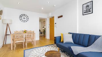 A living room with a blue sofa, a wooden dining table with chairs, a round coffee table, and a wall clock.