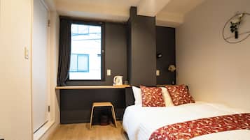 A hotel room with a bed, a small stool, a window with curtains, a kettle, and a wall-mounted light fixture.