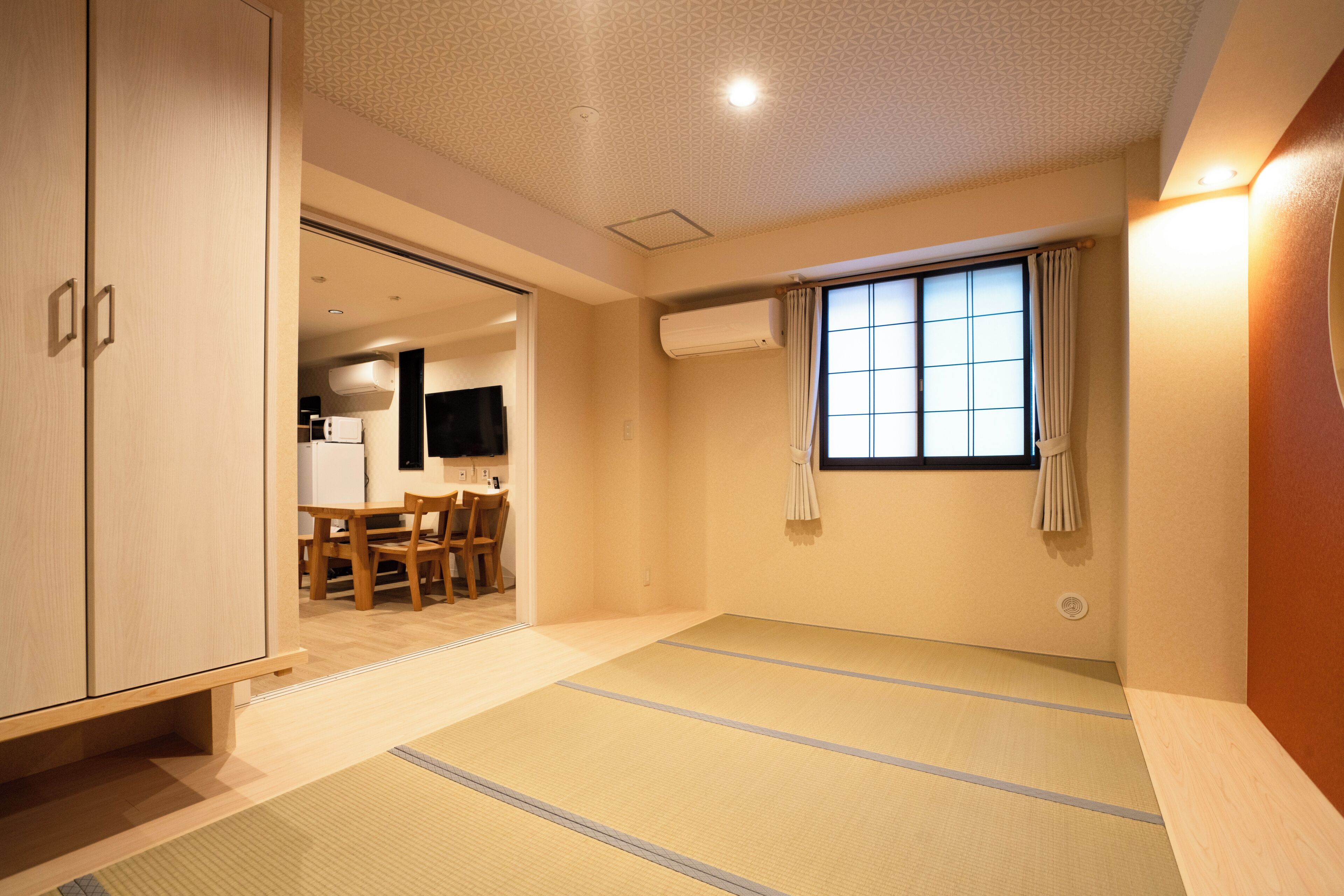 A room with tatami flooring, a sliding door, a window with curtains, and a kitchenette in the background.