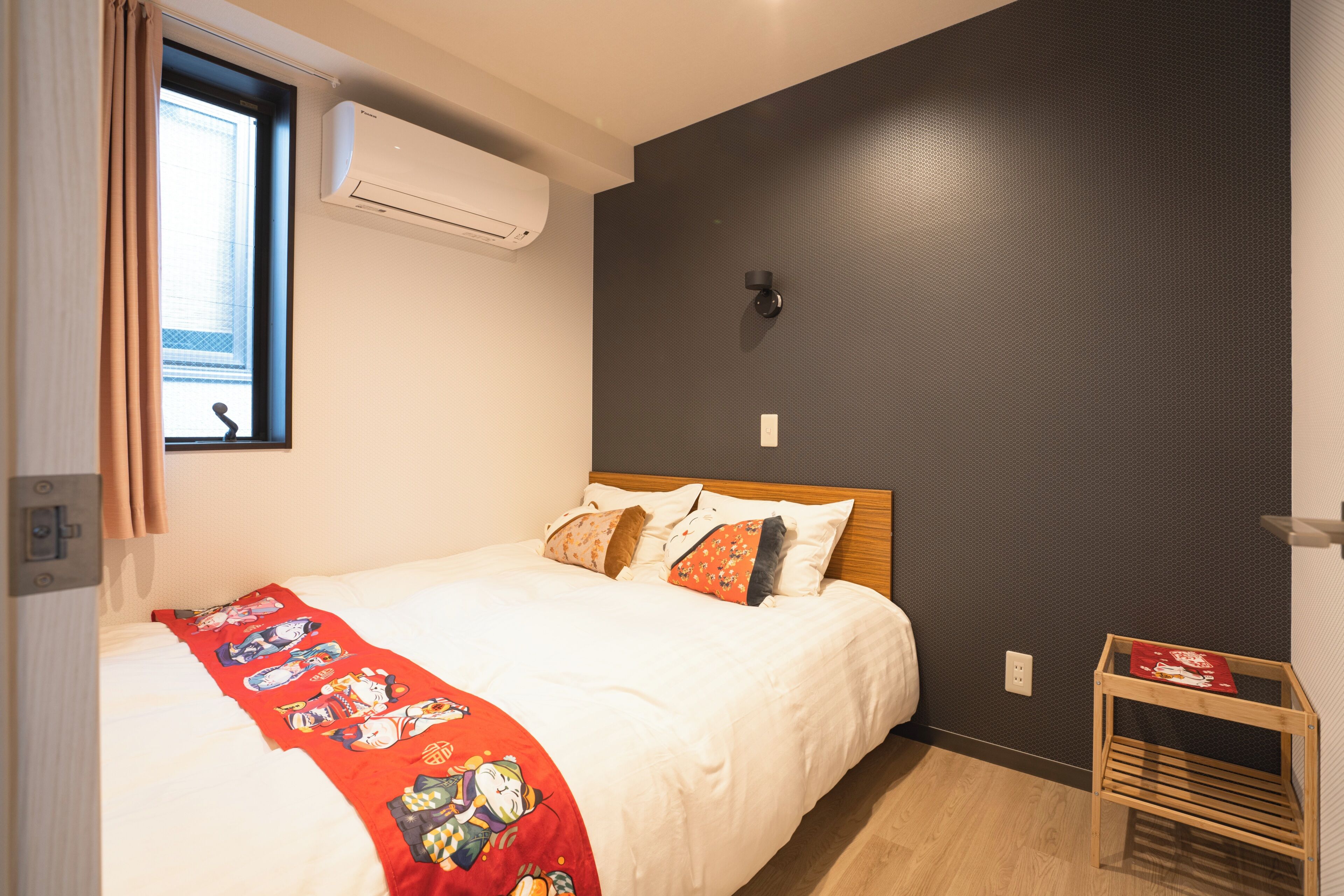 A bedroom with a bed, a window with curtains, an air conditioning unit, a wall-mounted light, and a small wooden table.