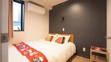 A bedroom with a bed, a window with curtains, an air conditioning unit, a wall-mounted light, and a small wooden table.