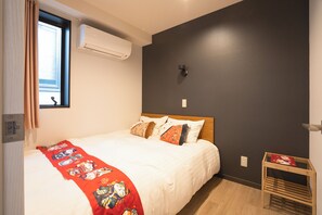 A bedroom with a bed, a window with curtains, an air conditioning unit, a wall-mounted light, and a small wooden table. - Tokyo Biyori Hotel (Tokyo)