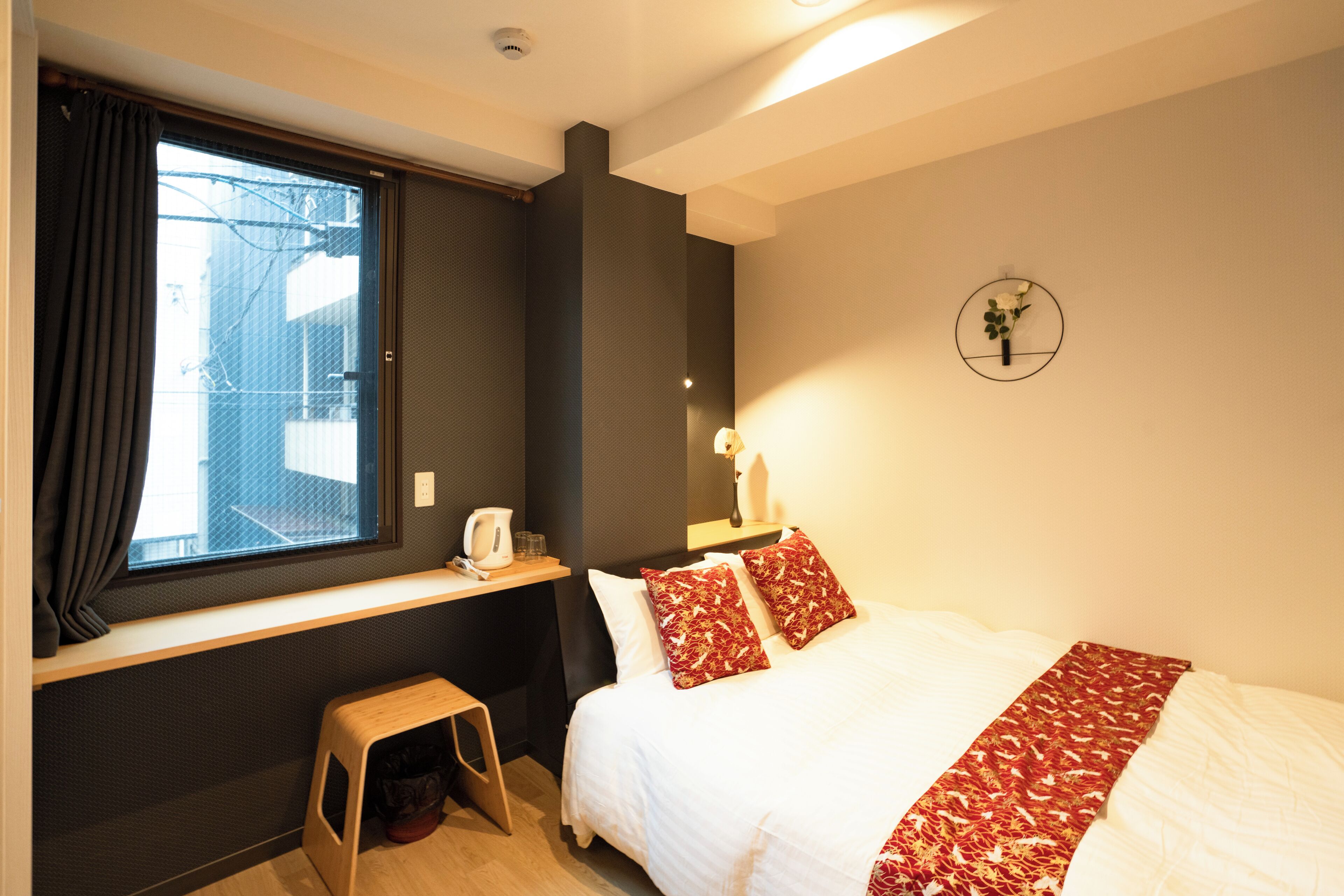 A hotel room with a bed, a window with curtains, a small stool, a bedside table with a kettle, and a wall-mounted clock.