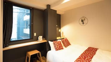 A hotel room with a bed, a window with curtains, a small stool, a bedside table with a kettle, and a wall-mounted clock.