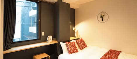 Una habitación de hotel con una cama, un ventanal con cortinas, un taburete pequeño, una mesita de noche con hervidor y un reloj de pared.