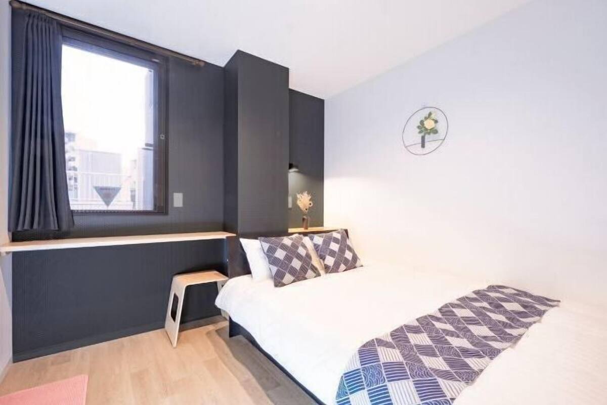 A bedroom with a bed, a window with curtains, a bedside table, and a wall-mounted light fixture.