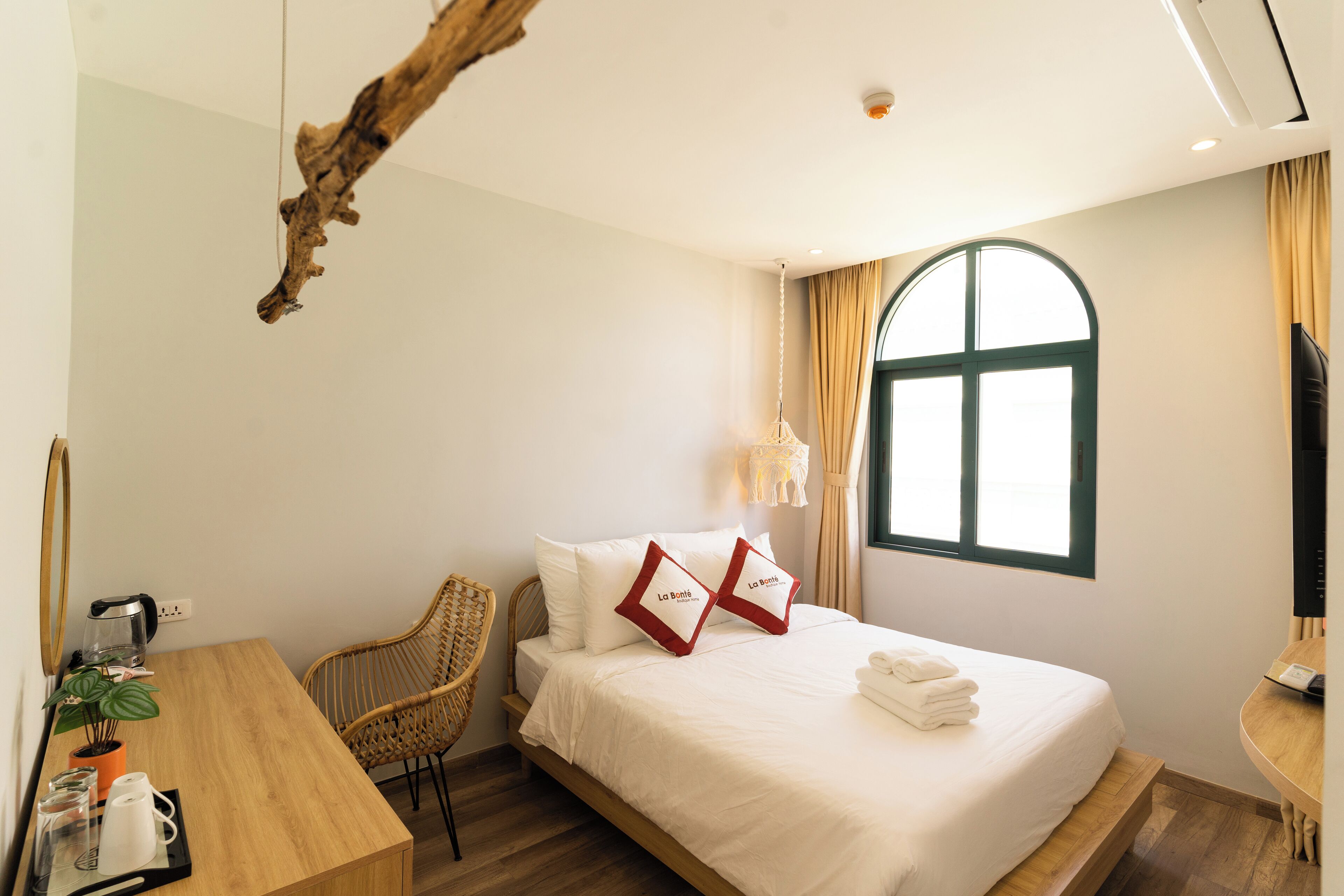 A hotel room with a bed, a wooden desk, a chair, a potted plant, and a window with curtains.