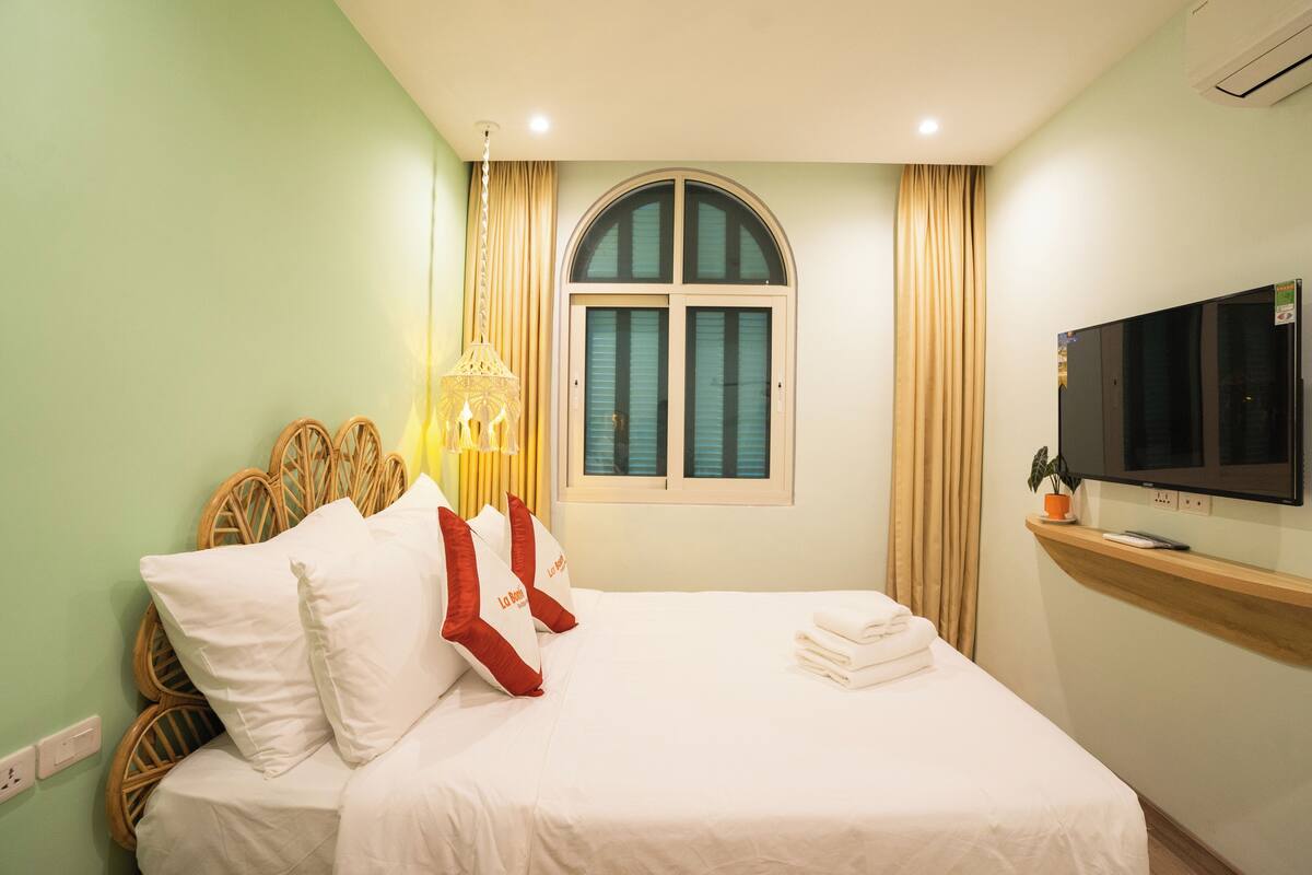 Une chambre d’hôtel avec un lit, une télévision, une fenêtre avec des rideaux et une étagère fixée au mur.