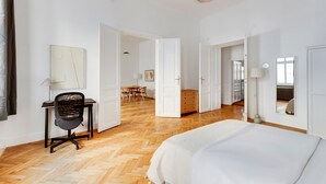 A spacious hotel room with a large bed, a desk with a chair, and a dining area in the background.