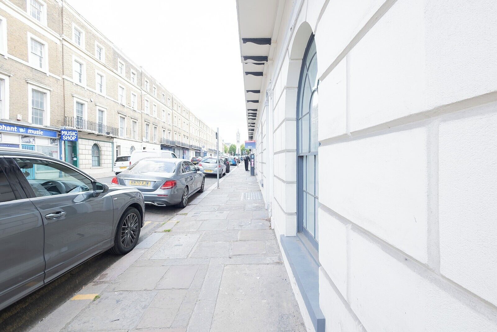 A narrow street with parked cars and a building with a white facade and large windows.