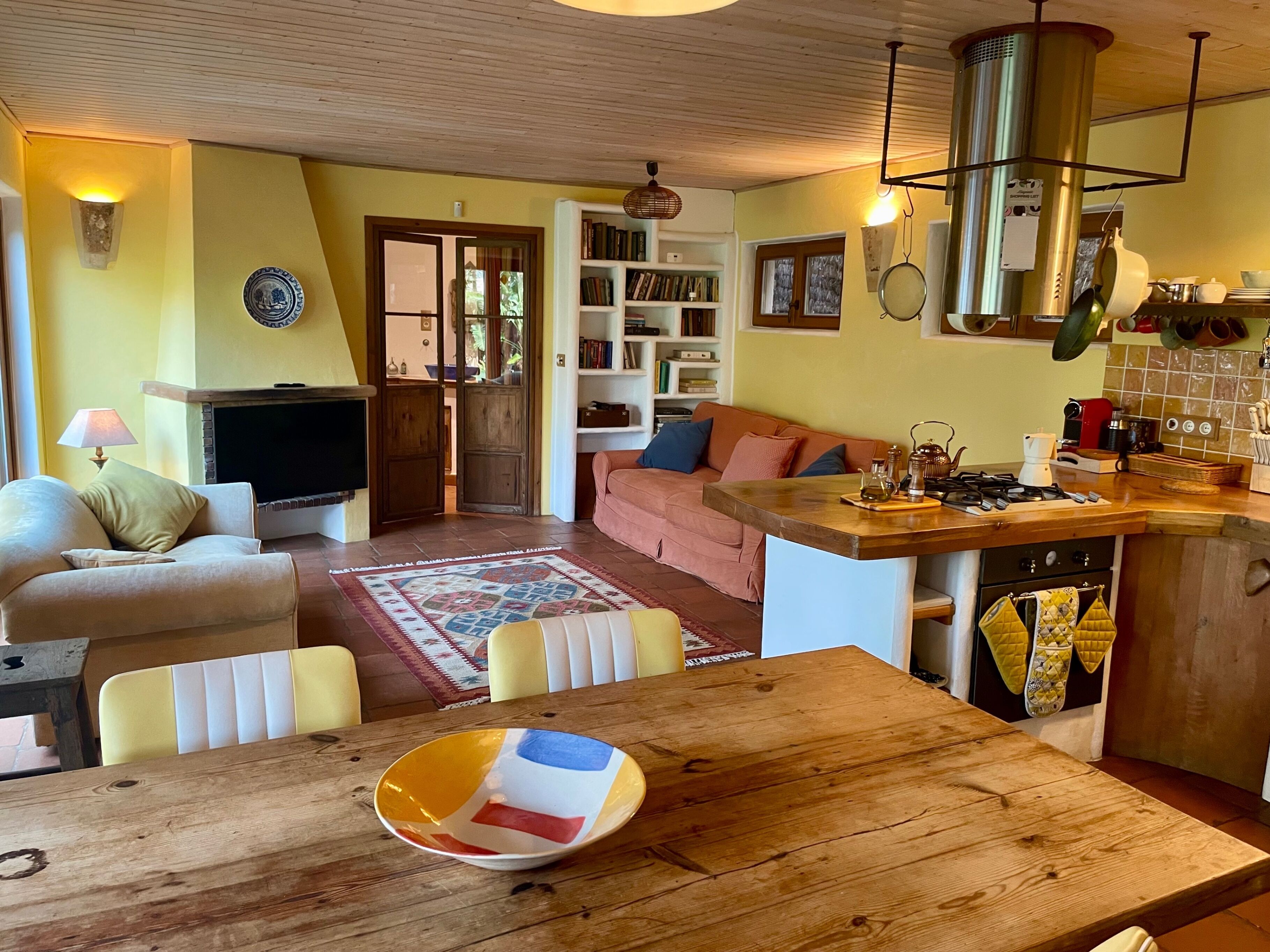 Un salon chaleureux avec une cheminée, un canapé, une table à manger avec un saladier, et un coin cuisine équipé d’une cuisinière et d’ustensiles.