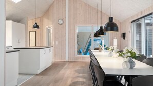 Eine moderne Küche mit Holzvertäfelung, ein Essbereich mit einem weißen Tisch und schwarzen Stühlen sowie Blick auf ein blaues Schwimmbad durch eine Glastür.