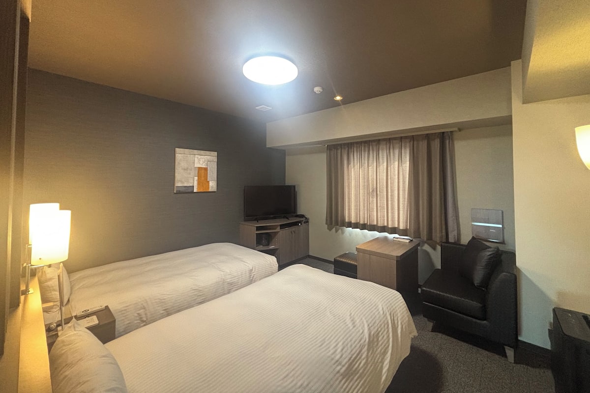 Une chambre d’hôtel avec deux lits, un bureau, une chaise, une télévision et une fenêtre avec des rideaux.