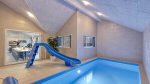 Una piscina interior con un tobogán azul, un área de comedor con mesa y sillas, y una planta en maceta.