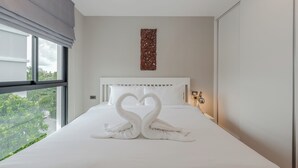 A modern hotel room with a large bed, a view of greenery outside, and a decorative wall piece above the bed. - Modern Condo Walk to Rawai Beach C188 (Rawai)