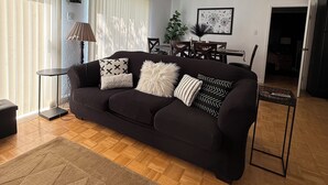 A living room with a black sofa, a wooden floor, a ceiling fan, and a dining table with chairs.