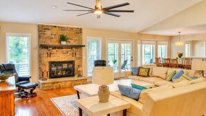 A spacious living room with a stone fireplace, a ceiling fan, and a large window. - Pool House on Ponce De Leon (Hot Springs)