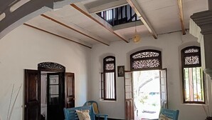 A room with a high ceiling, wooden beams, and a large window with intricate lattice design. There are two blue chairs and a table with a cushion. - The Dutch Inn (Galle)