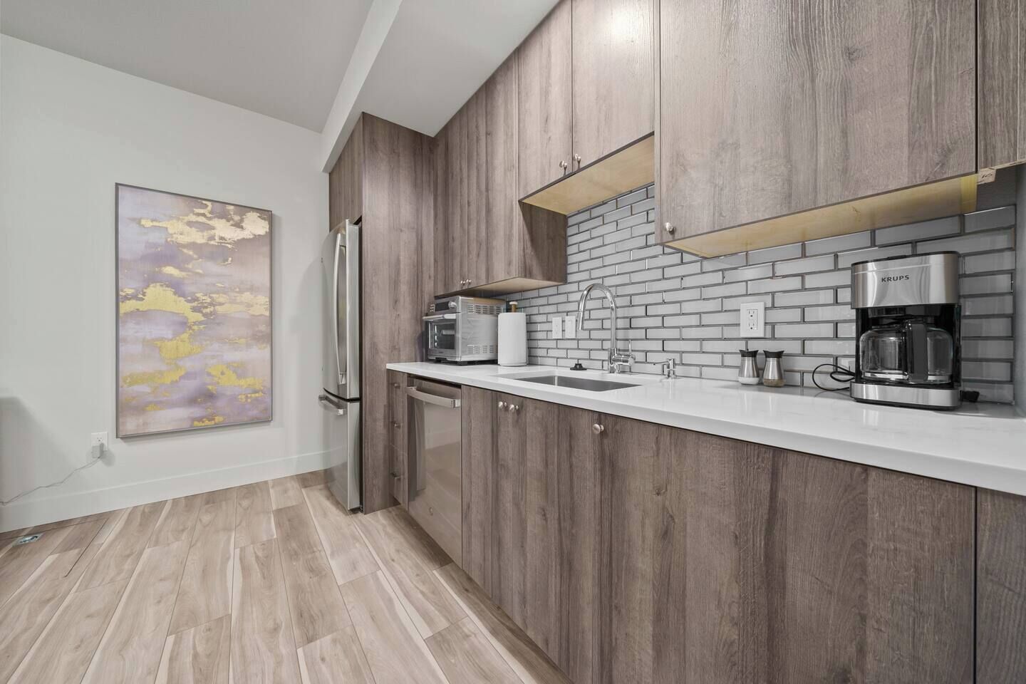 A modern kitchen with wooden cabinets, a white countertop, a stainless steel refrigerator, a microwave, a coffee maker, and a brick backsplash.
