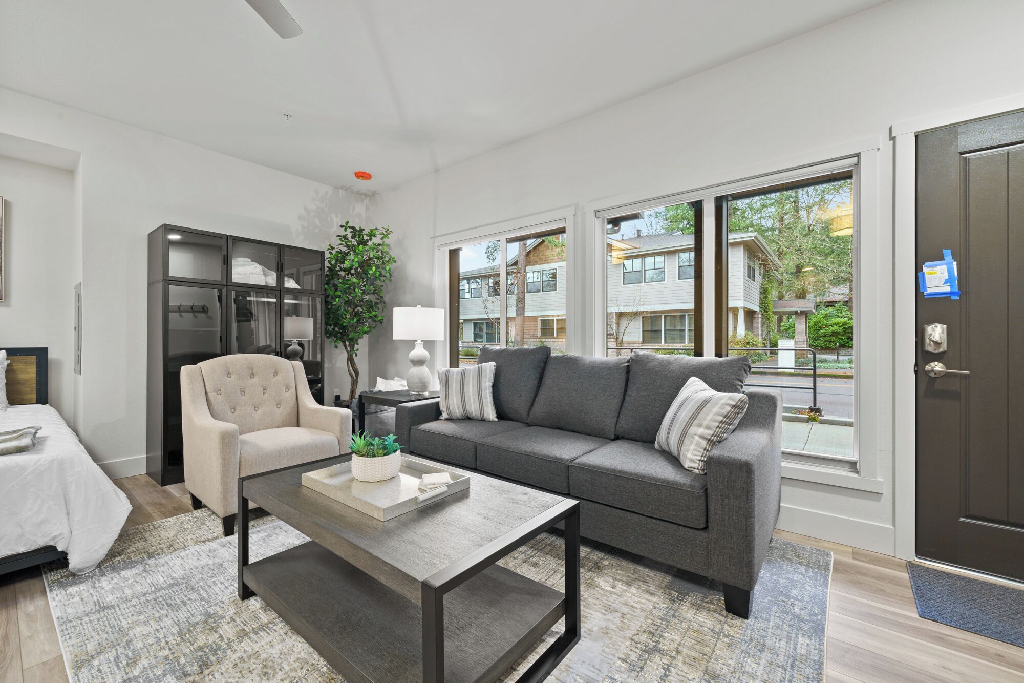 A modern living room with a sofa, armchair, coffee table, and a large window with a view of the neighborhood.