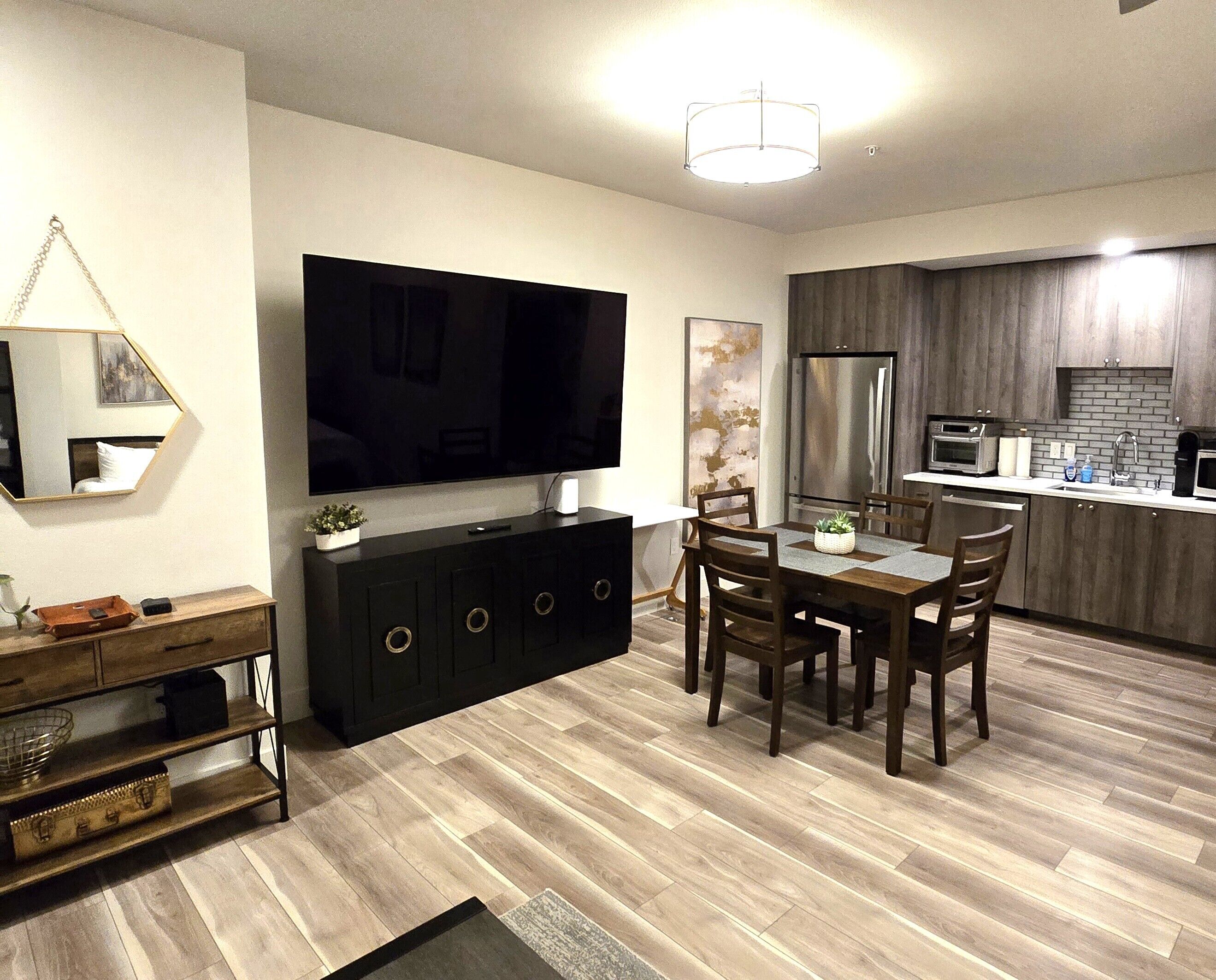 A modern living room with a flat-screen TV, dark wooden dining table, and chairs. A kitchen area with a refrigerator and microwave is visible in the background.