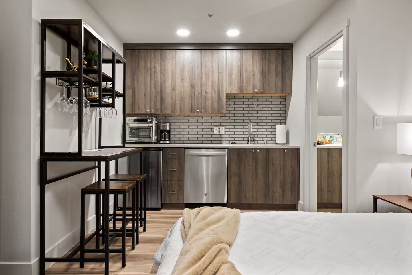 A compact kitchen with wooden cabinets, a built-in oven, and a dishwasher. There is a small bar area with stools and a bed with a blanket in the foreground.