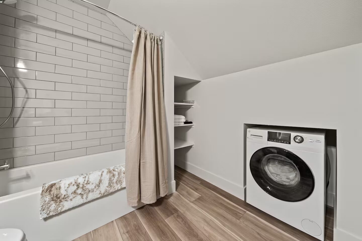 A modern bathroom with a white tiled wall, a wooden floor, a bathtub, a washing machine, and a beige curtain.