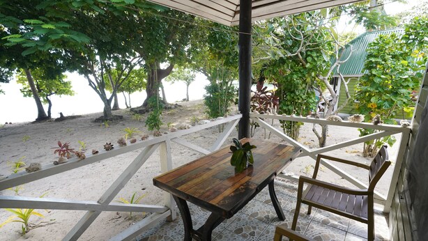 A wooden table with a potted plant, surrounded by a white railing and a bench, set on a tiled patio with a view of a beach and trees. - White Sandy Beach Eco Lodge (Naviti Island)