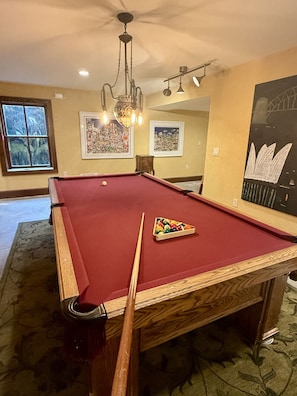 A pool table with a cue stick, a chandelier, framed artwork on the walls, and a window with a view.