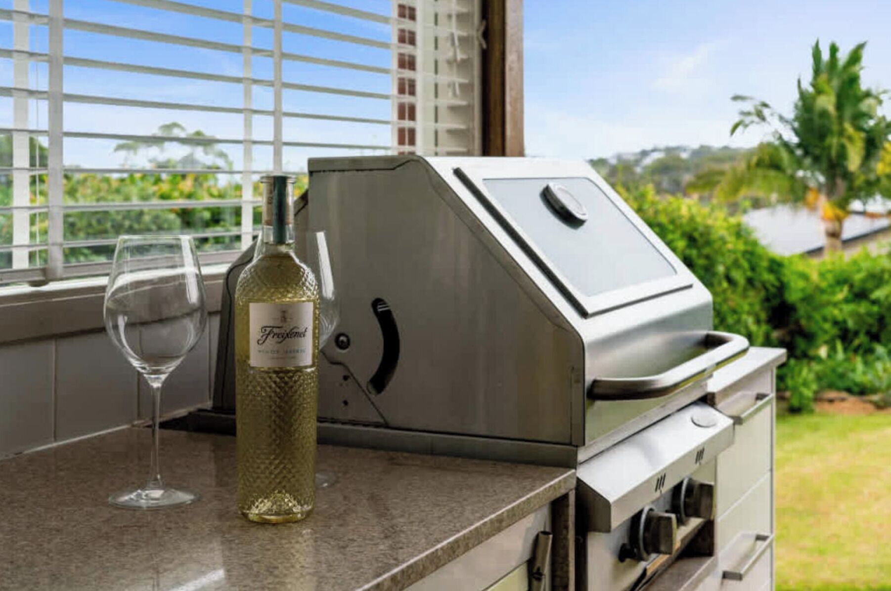 A stainless steel grill with a wine bottle and glass on a countertop near a window with blinds.