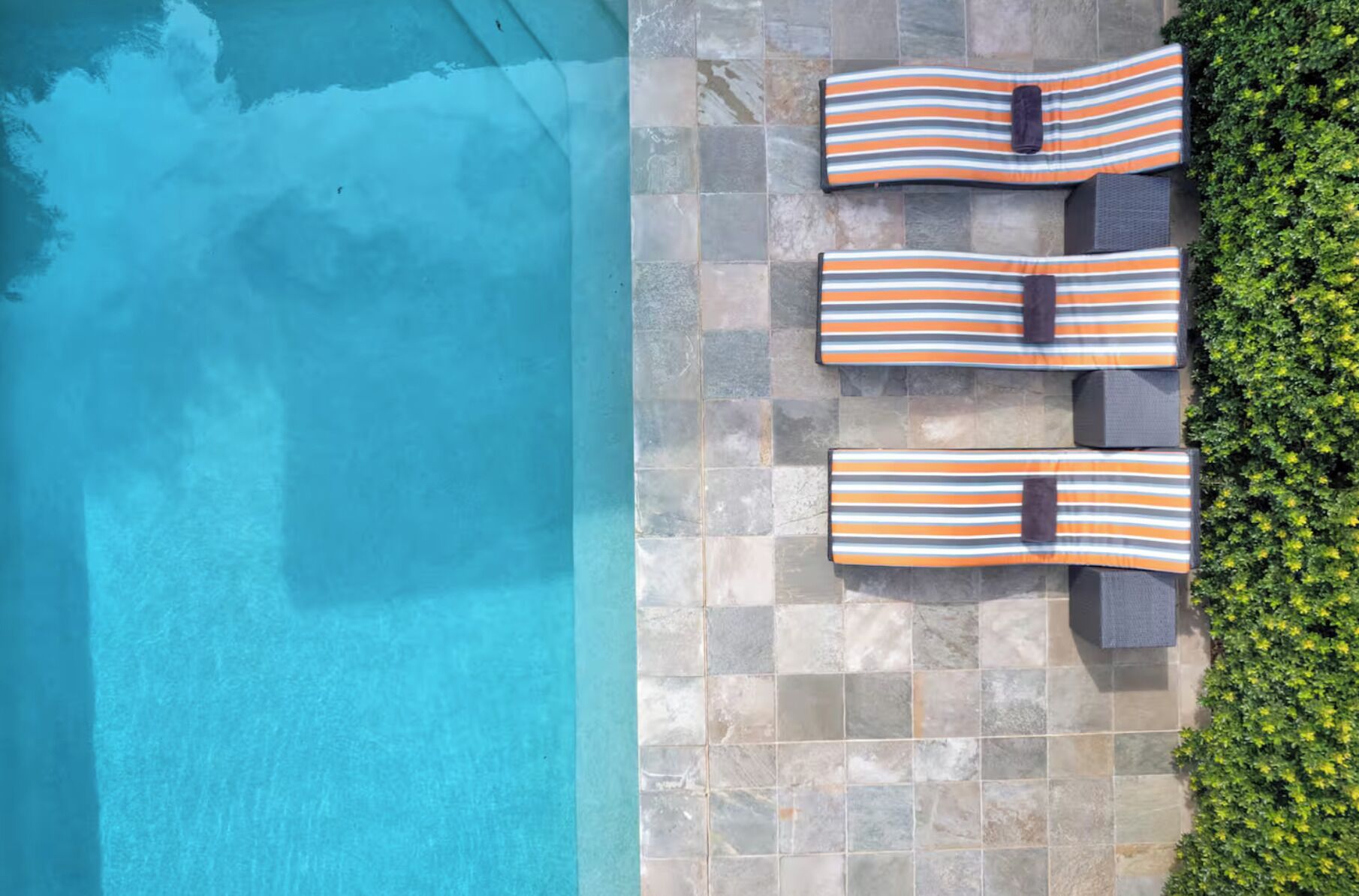 An aerial view of a swimming pool with three lounge chairs and a stone tiled deck.