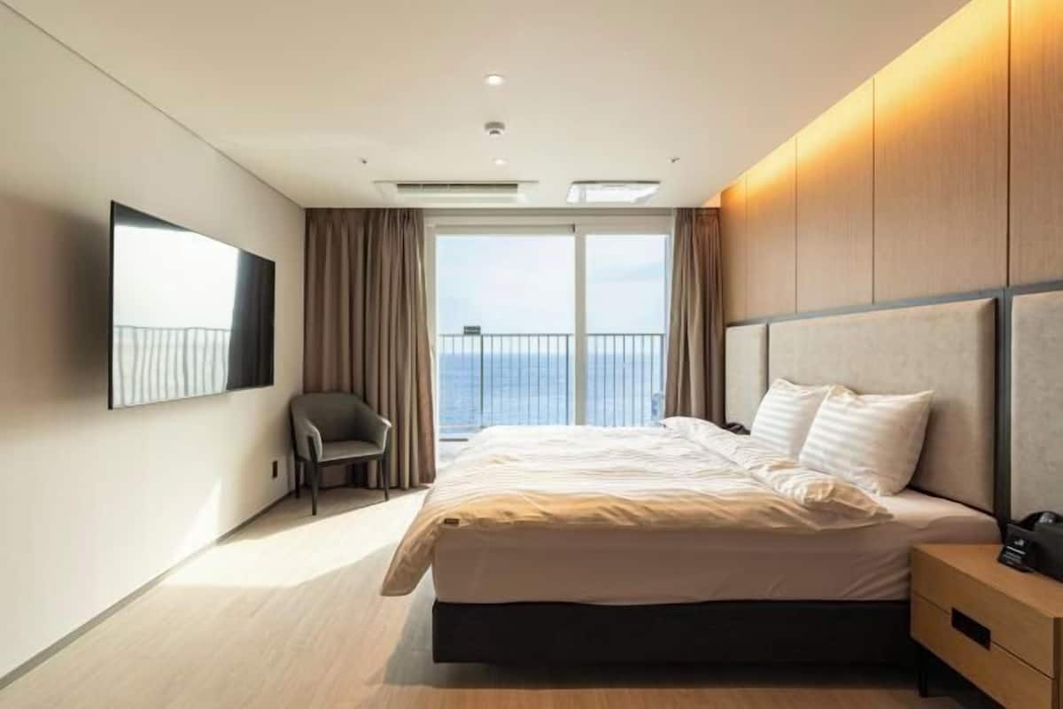 A modern hotel room with a large bed, a chair, a TV, and a balcony with a view.