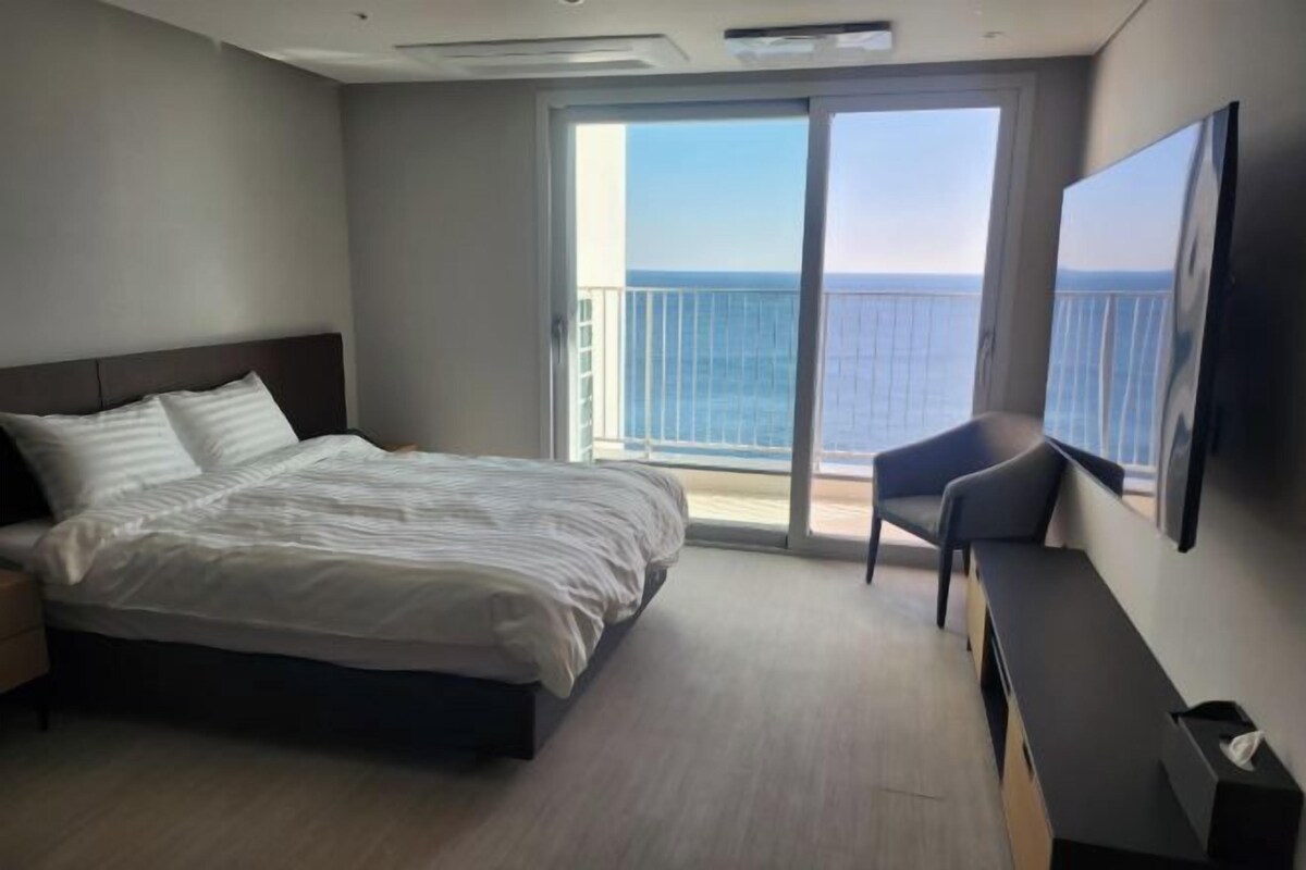 A bedroom with a bed, a chair, a TV, and a balcony with a view of the sea.