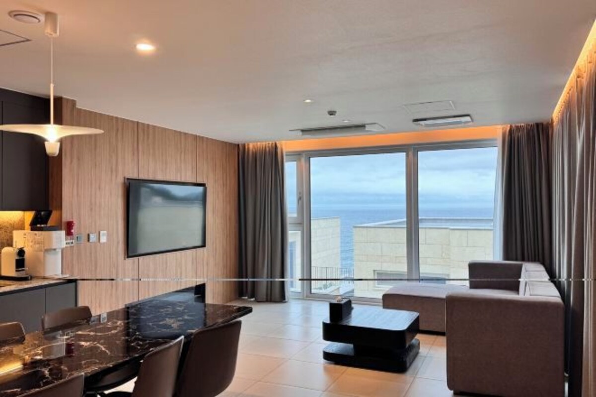 A modern hotel room with a kitchen area, a dining table, a sofa, and a large window offering a view of the sky and buildings.
